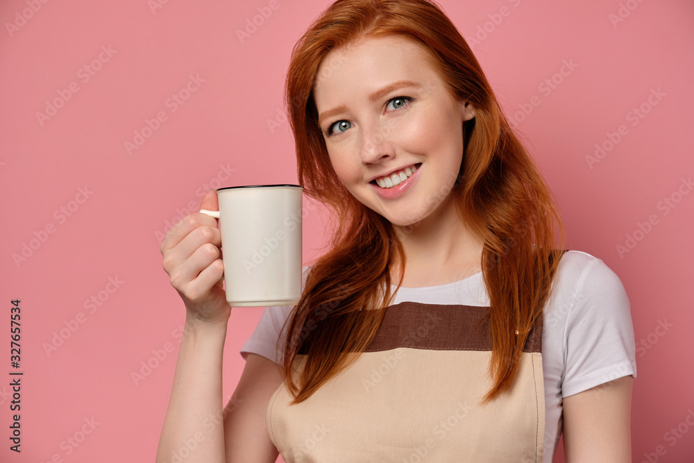 Beautiful red-haired girl in an apron stands on a pink background and smiles, holding a white mug to her face.