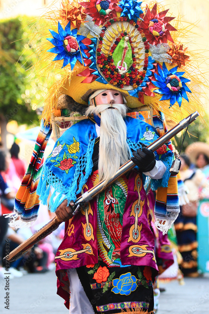 a mexican carnival dancer with colorful costume, icon of Our Lady of Guadalupe as part of the decoration holds a gun on his hands