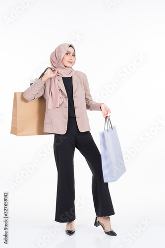 A beautiful Asian Muslim woman wearing officewear and hijab poses with shopping bags isolated on white background. Full length portrait for business and commerce concepts.