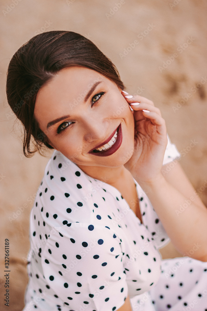 Portrait of cheerful beautiful brunette girl in white polka dot dress sitting on sandy beach and smiling. Young cute woman in stylish retro clothing posing at seaside. Beach after party outdoors Portrait of cheerful beautiful brunette girl in white polka dot dress sitting on sandy beach and smiling. Young cute woman in stylish retro clothing posing at seaside. Beach after party outdoors
