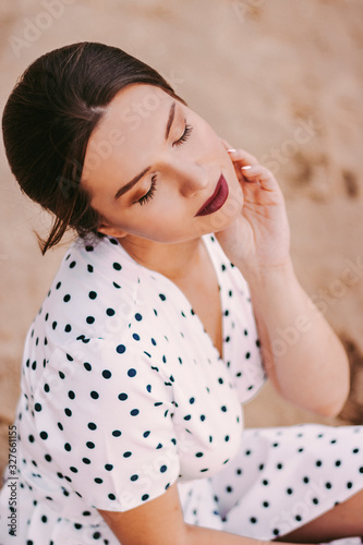 Portrait young tender brunette girl in white polka dot dress keeping eyes closed and smiling while sitting on sandy beach. Beautiful happy woman in stylish retro clothing posing at seaside after party