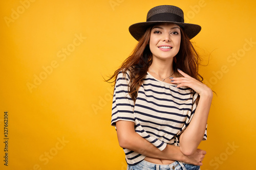 A beautiful brunette in a striped T-shirt and wicker hat stands on a yellow background, smotot into the frame,raising hand to neck