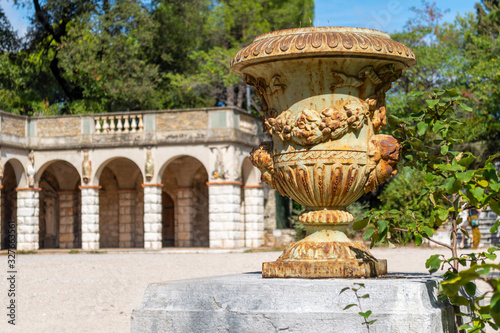 An ornate antique vase sits atop a column with a historic arched building blurred behind on Castle Hill in Nice, France.
