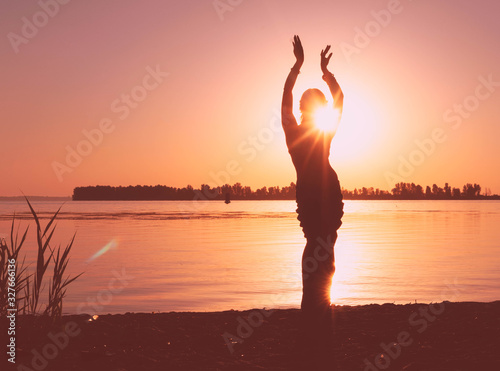 glowing silhouette of slim woman with hands up in the air illuminated with sunshine in front sky near river at sunrise