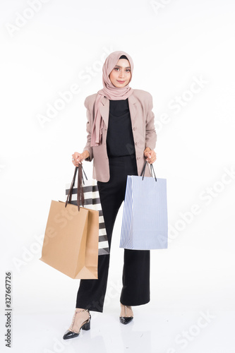 A beautiful Asian Muslim woman wearing officewear and hijab poses with shopping bags isolated on white background. Full length portrait for business and commerce concepts.