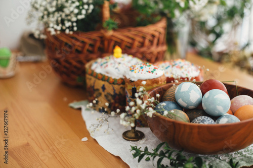 фотографія Stylish easter eggs natural dyed on background of homemade easter cake, candle, green branches and flowers with wicker basket on wooden rustic table