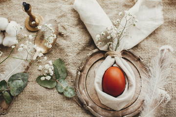 Stylish Easter flat lay, table setting with egg in easter bunny napkin. Modern natural dyed egg on napkin with bunny ears, flowers on vintage plate. Rustic Easter table decorations