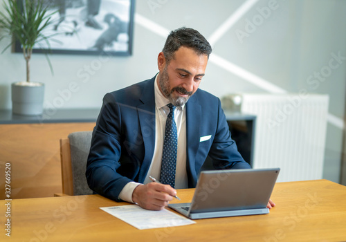 The boss, who checks and takes notes at the computer in his own workroom in the office, the handsome hardworking boss with the beard in the suit, the bank manager, the team leader