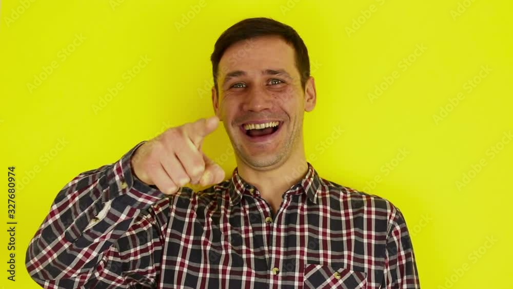 A young handsome guy in a plaid shirt looks at the camera, laughs expressively and points a finger at the interlocutor. Portrait on a yellow background.