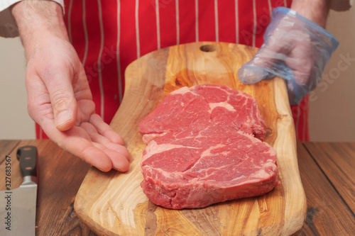 Butcher in red apron holding wooden board with two fresh rib eye steaks with his left hand in plastic glove, right hand pointing to the steaks, knife lays on the wooden table. Meat industry concept.