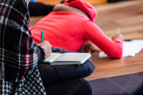 The unrecognizable lady who takes notes on her notebook, while doing yoga among other people who do practicing yoga and making notes as she makes