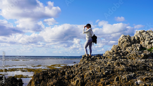 Young girl traveler with a backpack on her shoulders stands on top of a rocky mountain on the Pacific Ocean at low tide. Sea view on the Japanese island of Ishigaki in Okinawa. Background of clear sky