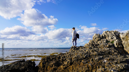 A young girl in gray sportswear and a black cap is standing on the rocky shore of the Pacific Ocean and looks at the stone coast at low tide. Sea view on the Japanese island of Ishigaki in Okinawa