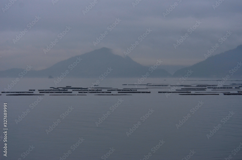 A bay at sunset, filled with mist. Mountains are on the far shore. Wooden floats used for fishing and oyster farming are in the foreground.