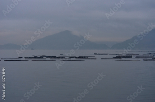 A bay at sunset, filled with mist. Mountains are on the far shore. Wooden floats used for fishing and oyster farming are in the foreground.