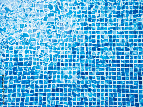 The light reflected on the top of the water surface of the pool lined with blue mosaic tiles below. Water ripples on blue-tiled swimming pool background.