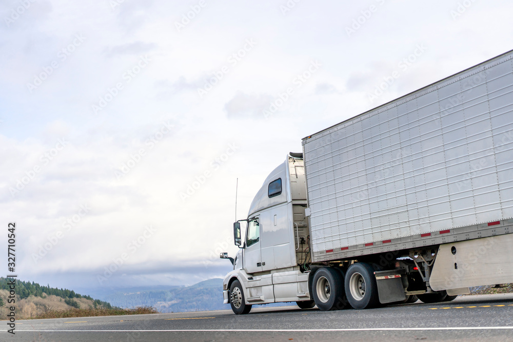 White big rig semi truck transporting food in refrigerator semi trailer driving on the road with sky and hills view White big rig semi truck transporting food in refrigerator semi trailer driving on the road with sky and hills view