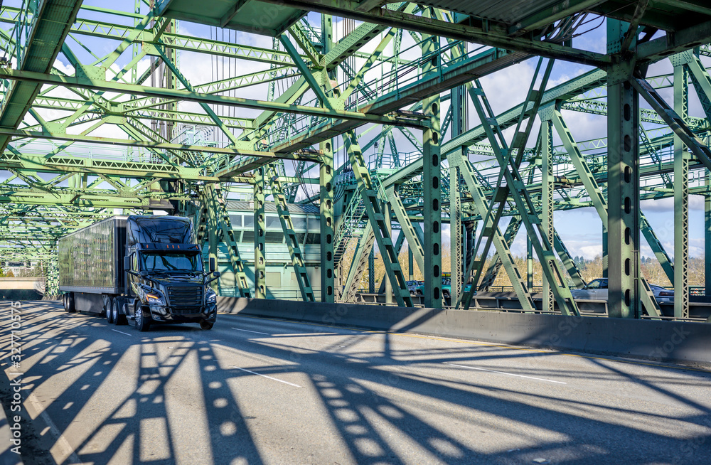 Fototapeta premium Stylish black big rig semi truck with covered black semi trailer running with cargo on the truss bascule Columbia River Interstate Bridge in sunny day