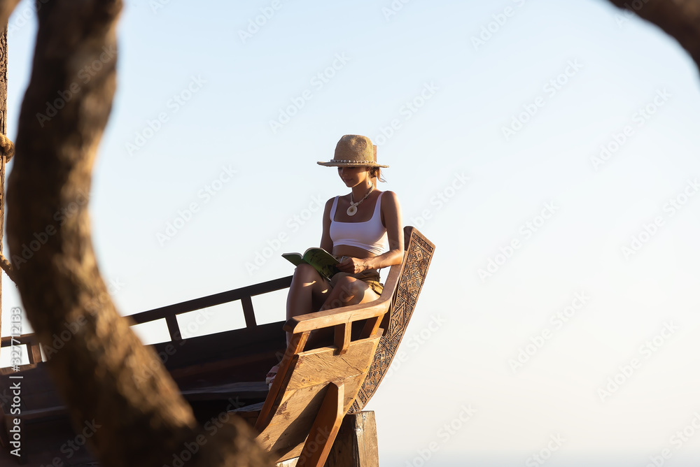 Young woman reading book and looking at beautiful sunrise on the pier with sea and mountains on background Young woman reading book and looking at beautiful sunrise on the pier with sea and mountains on background