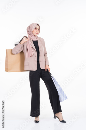 A beautiful Asian Muslim woman wearing officewear and hijab poses with shopping bags isolated on white background. Full length portrait for business and commerce concepts.