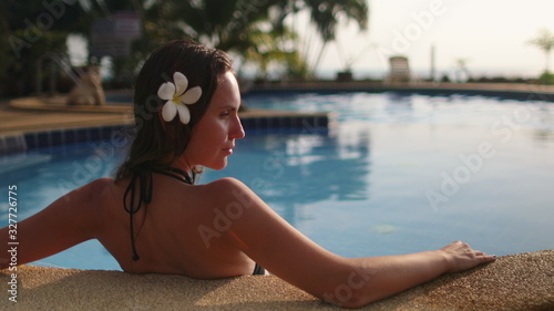 Caucasian Woman Sitting in Resort Swimming Pool. Young Model Sunbathing in Turquoise Water. Relaxed Girl with Exotic Flower in Brunette Hair Enjoying Koh Phangan Island.