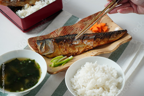 someone hand using chopsticks trying to pick a grilled saba or mackerel fish served with miso soup and cooked rice on white and green striped placemat on white table