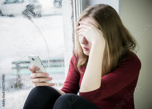A teenager girl with long blond hair sits by the window on a windowsill with a phone in her hand and covered her face with her hand. The concept of teenage addiction to a mobile phone, the Internet.