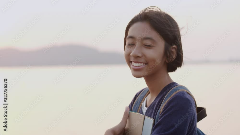 Happy Asian student smile on face while standing on the beach at the sunset or sunrise, Asia child hold book and backpack on sea background. The schoolgirl came to study field trip and learned outdoor Happy Asian student smile on face while standing on the beach at the sunset or sunrise, Asia child hold book and backpack on sea background. The schoolgirl came to study field trip and learned outdoor