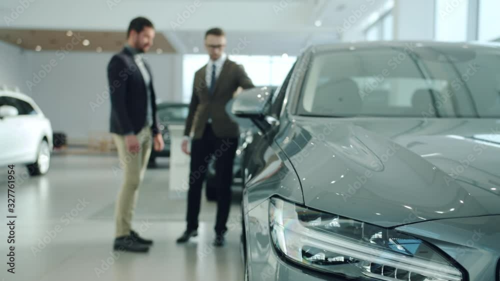 Handsome guy is talking to professional sales agent in car dealership, focus on expensive automobile. Transportation, luxury and modern vehicles concept. Focus on car headlight