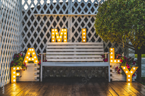 The word FAMILY is decorated with glowing letters. There is a bench between the letters . There is a wooden fence at the back and a green tree at the side. Day