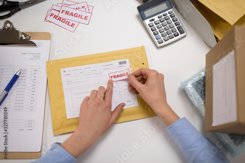 delivery, mail service, people and shipment concept - close up of woman's hands sticking fragile marks to parcel in envelope at post office