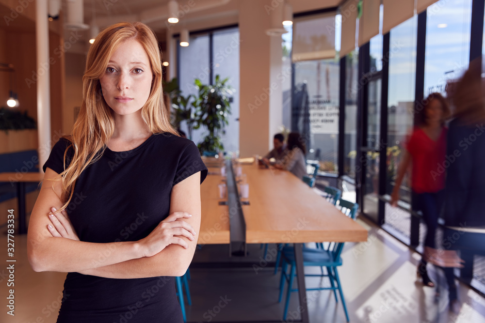 Portrait Of Businesswoman Standing In Busy Modern Open Plan Office  With Colleagues In Background