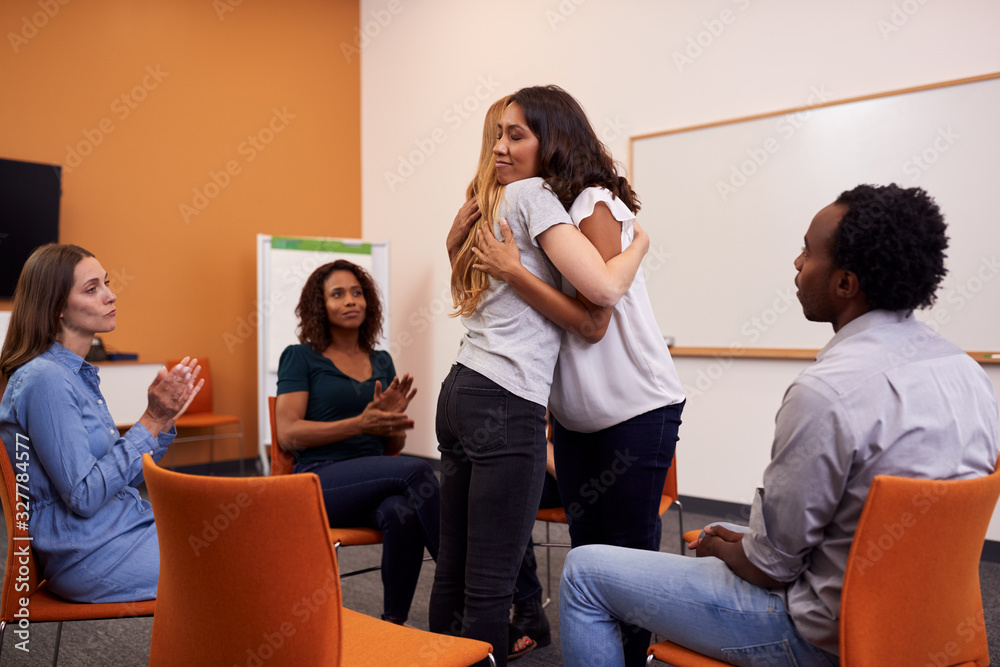 Two Women Hugging As Group Of Men And Women At Mental Health Group Therapy Meeting Two Women Hugging As Group Of Men And Women At Mental Health Group Therapy Meeting