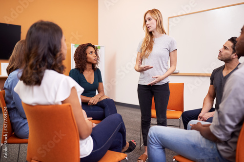 Woman Standing To Address Group Of Men And Women At Mental Health Group Therapy Meeting