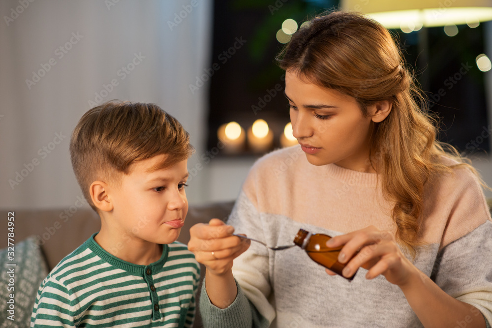 family, health and people concept - sad mother pouring antipyretic medication or cough syrup to spoon for ill little son at home family, health and people concept - sad mother pouring antipyretic medication or cough syrup to spoon for ill little son at home