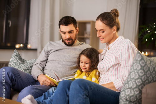 family, leisure and people concept - happy father, mother and little daughter reading book at home at night