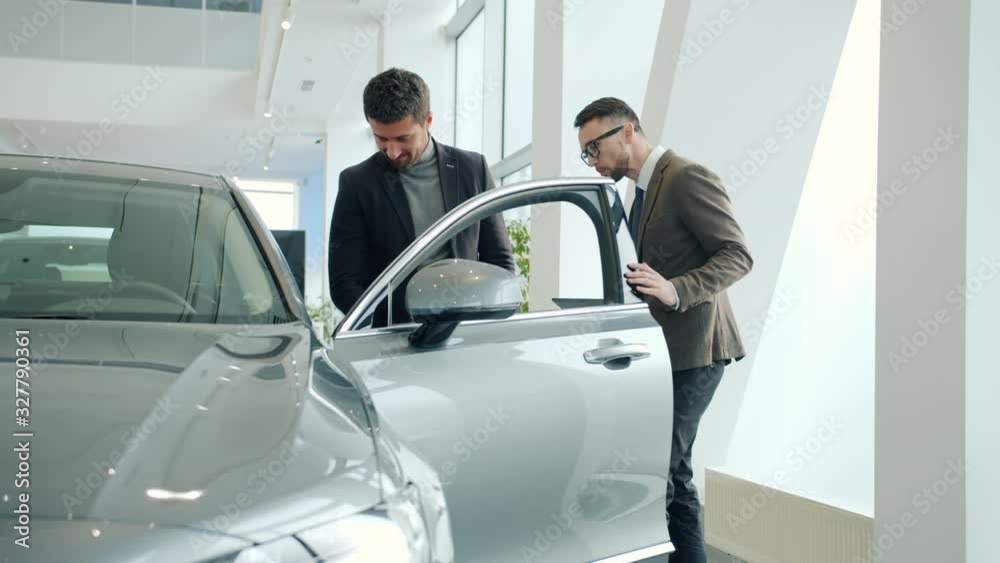 Salesman friendly young man with tablet is opening car door for buyer selling new automobile in dealership. Business, transport and conversation concept. Salesman friendly young man with tablet is opening car door for buyer selling new automobile in dealership. Business, transport and conversation concept.
