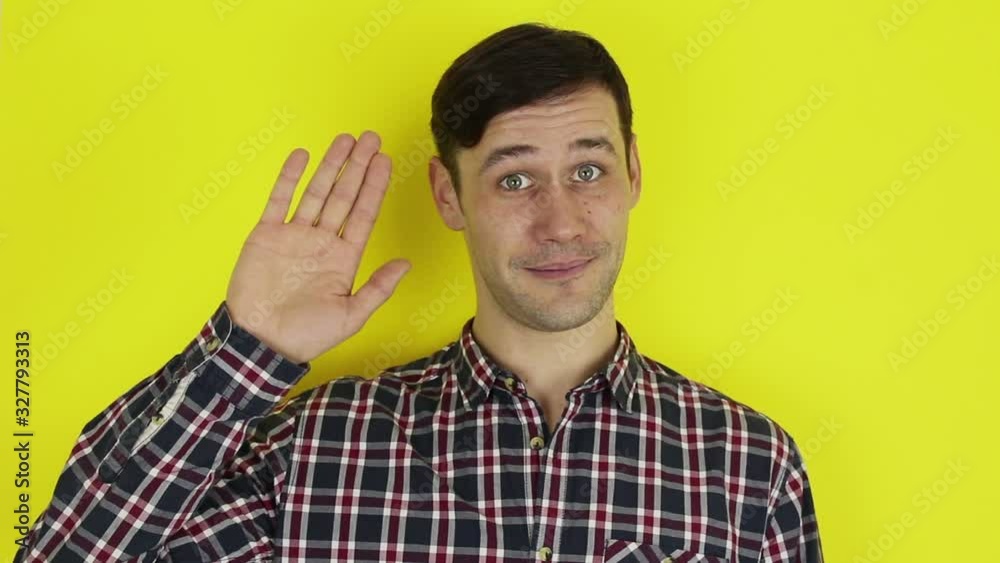 Young handsome guy in a plaid shirt, says goodbye and waves his hand .Portrait on a yellow background. Young handsome guy in a plaid shirt, says goodbye and waves his hand .Portrait on a yellow background.