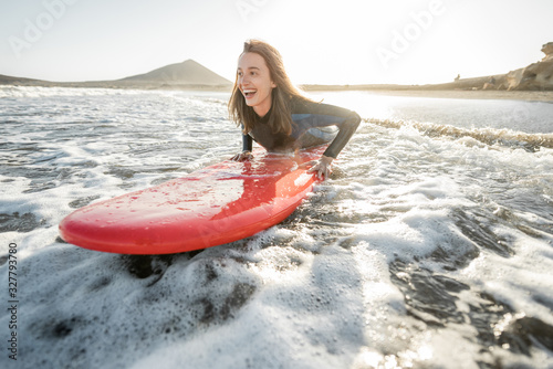 Photography Young woman in wetsuit catching water flow on the surfboard, surfing on the wavy ocean during a sunset