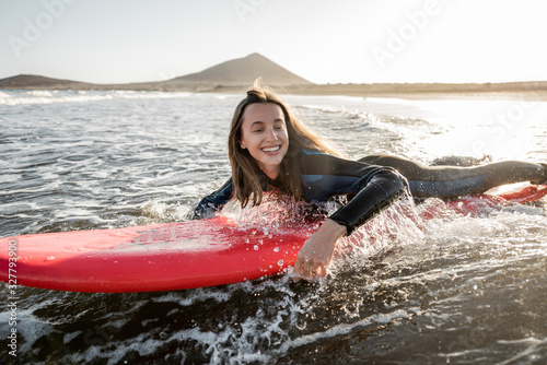 Photography Young woman in wetsuit catching water flow on the surfboard, surfing on the wavy ocean during a sunset