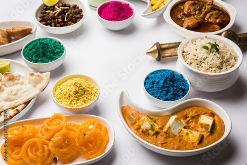 Happy Holy concept  showing Indian assorted lunch food like chicken  paneer butter masala  naan  jeera rice  black chana fry  jalebi  fujiyama  thandai and Farsan with holi colours and pichkari 