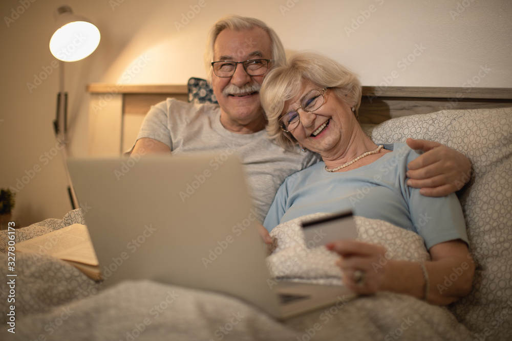 Cheerful senior couple e-banking on laptop while resting in bed.