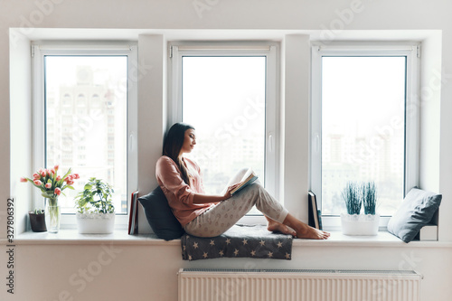 Fototapet Carefree young woman in cozy pajamas reading a book while resting on the window