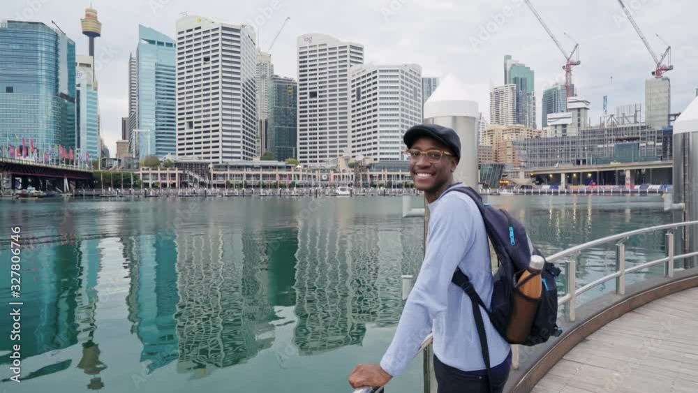 Black man observes and studies the Sydney skyline from the Darling Harbour sidewalk, then smiles back at the camera. Black man observes and studies the Sydney skyline from the Darling Harbour sidewalk, then smiles back at the camera.
