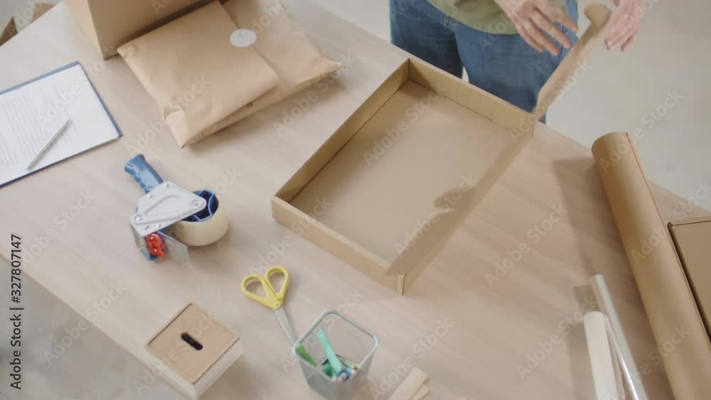 Flat lay of unrecognizable online shop worker standing at wooden table and putting ordered good wrapped in craft paper into box