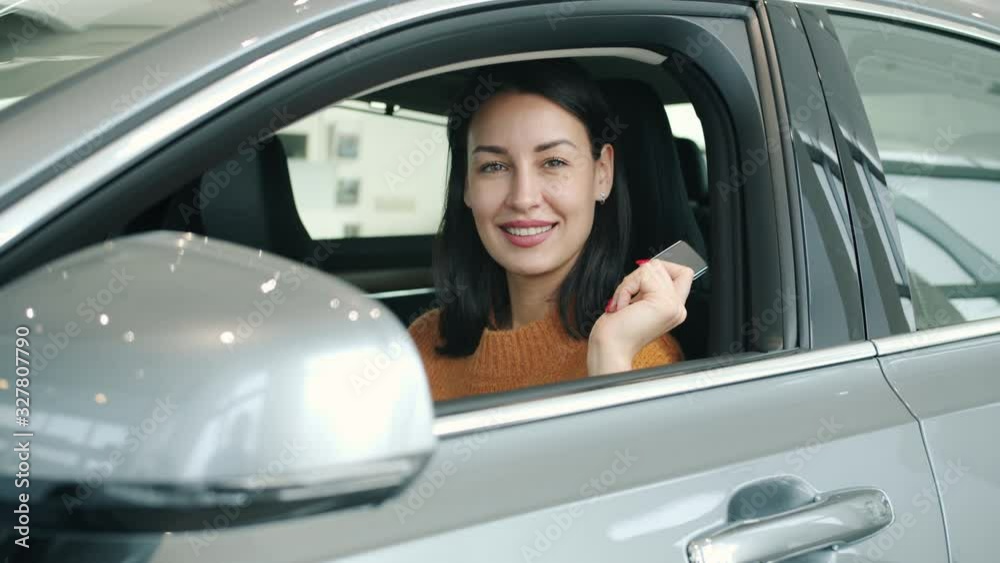 Slow motion portrait of pretty girl smiling sitting inside new car holding keys indoors in dealership. Positive emotions, transport and modern business concept. Slow motion portrait of pretty girl smiling sitting inside new car holding keys indoors in dealership. Positive emotions, transport and modern business concept.