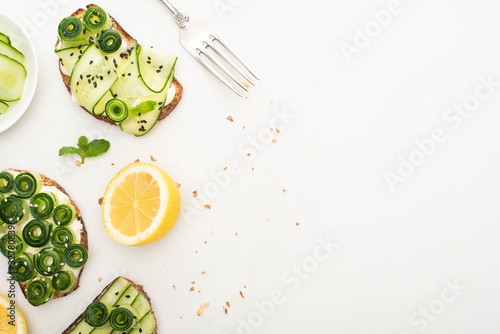 top view of fresh cucumber toasts with seeds, mint leaves near lemon and fork on white background