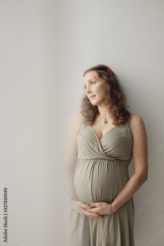 Portrait of pregnant woman standing near the window and touching her big belly. Waiting for the baby. Motherhood.