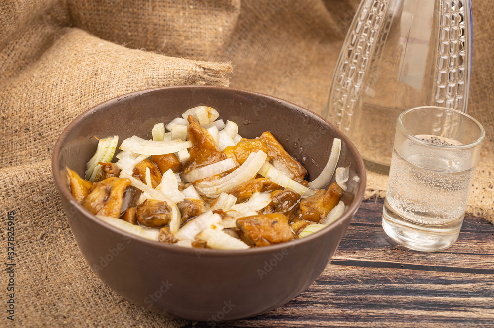 Salted mushrooms with onions in a ceramic bowl, a glass of vodka and a decanter on a background of rough homespun fabric. Country food. Close up. Salted mushrooms with onions in a ceramic bowl, a glass of vodka and a decanter on a background of rough homespun fabric. Country food. Close up.