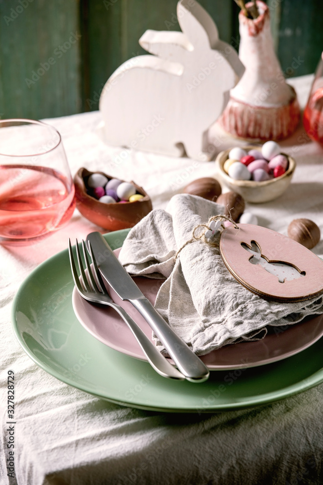 Easter table setting with wooden rabbit decoration, chocolate eggs and sweets, glasses of pink cocktail drinks, empty plates with textile napkin, cutlery on white tablecloth. Holiday dinner. Close up Easter table setting with wooden rabbit decoration, chocolate eggs and sweets, glasses of pink cocktail drinks, empty plates with textile napkin, cutlery on white tablecloth. Holiday dinner. Close up